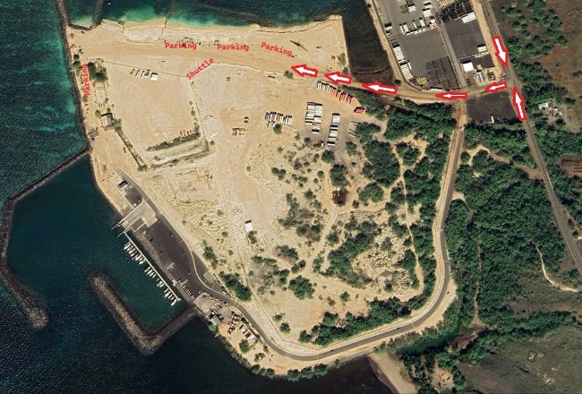 Aerial satellite view of a coastal industrial and harbor area, overlaid with red text and white arrows indicating a traffic route and parking locations. From a main road on the right, white arrows point west along a dirt path leading into a large, sandy clearing. Red labels mark several "Parking" zones along the northern and western edges of the clearing, and one "Shuttle" location near the center. The surrounding area includes a marina with docked boats to the southwest, a storage facility with stacked materials to the northeast, and green vegetation bordering the paved access road.
